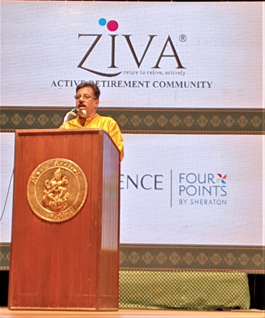 Dr. Sundararaman delivering introductory speech at podium with ZIVA Active Retirement Community and Four Points by Sheraton branding at Music Academy